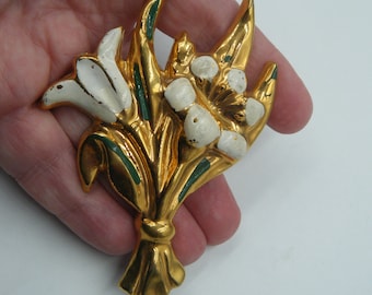 May include: A gold-tone metal brooch shaped like a bouquet of flowers. The design features polished gold stems and leaves with green enamel accents. The flower petals are finished with white enamel, showing some wear and texture on the surface.