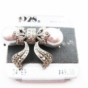 May include: A silver-toned brooch shaped like a bow, featuring two large, smooth, grey faux pearls on the sides and intricate, dark, faceted stone accents throughout the center and dangling ribbon ends. The brand name 1928 is printed above.
