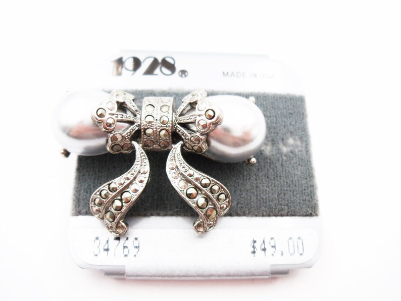 May include: A silver-toned brooch shaped like a bow, featuring two large, smooth, grey faux pearls on the sides and intricate, dark, faceted stone accents throughout the center and dangling ribbon ends. The brand name 1928 is printed above.
