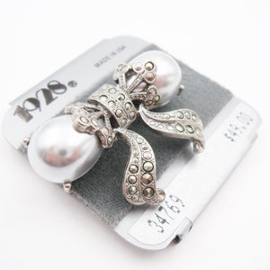 May include: A silver-toned brooch featuring two large, smooth, white faux pearls accented with dark, faceted marcasite stones arranged in a bow-like design. The piece is mounted on a gray display card labeled with the brand name 1928.
