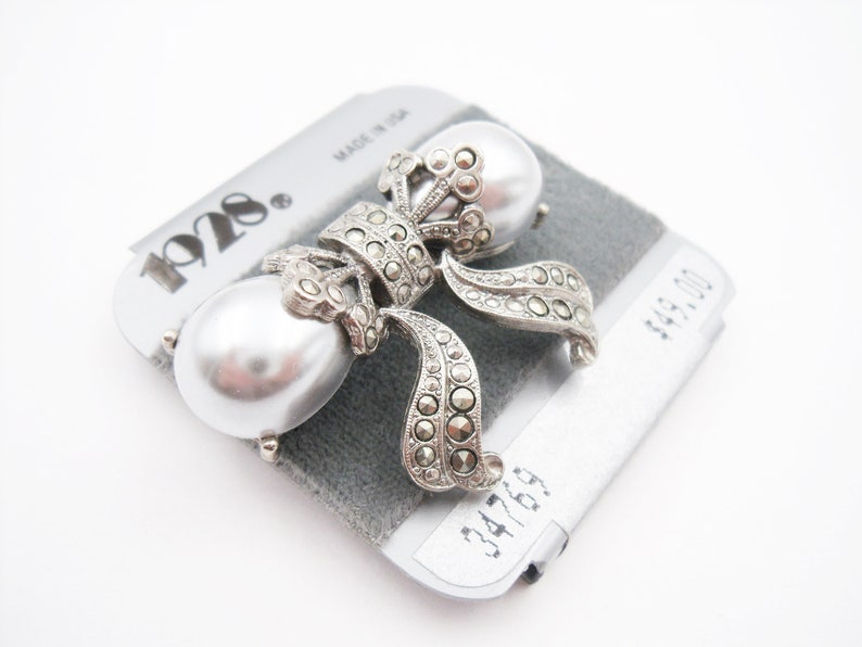 May include: A silver-toned brooch featuring two large, smooth, white faux pearls accented with dark, faceted marcasite stones arranged in a bow-like design. The piece is mounted on a gray display card labeled with the brand name 1928.