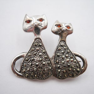 May include: A silver-toned cat brooch featuring two stylized cats side-by-side. The cats have orange gemstone eyes and bodies encrusted with small, dark, round stones. The brooch has a pin closure on the back.