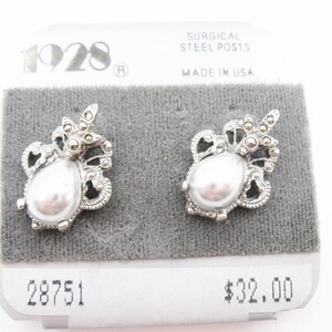 May include: A pair of silver-tone stud earrings featuring a central teardrop-shaped faux pearl surrounded by intricate metal filigree and small dark grey stones. The earrings are attached to a display card labeled 1928 and Surgical Steel Posts.