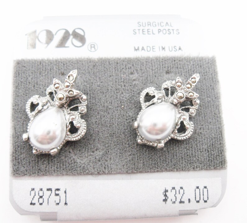 May include: A pair of silver-tone stud earrings featuring a central teardrop-shaped faux pearl surrounded by intricate metal filigree and small dark grey stones. The earrings are attached to a display card labeled 1928 and Surgical Steel Posts.