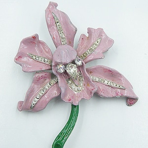May include: A vintage pink flower brooch with rhinestone detailing. The brooch has a green stem and a central cluster of rhinestones. The petals are a faded pink colour, adding to its antique charm.