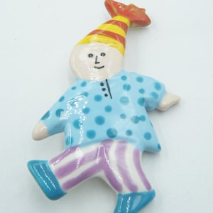 May include: A ceramic figurine of a whimsical clown. The clown wears a yellow and red striped hat, a light blue shirt with blue polka dots, and purple and white striped pants. The clown's shoes are blue.