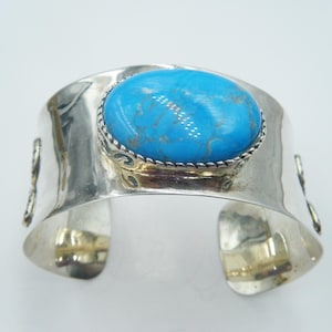 May include: A wide silver cuff bracelet featuring a large, oval turquoise stone set in a decorative bezel. The turquoise is a bright blue colour with darker veining. The bracelet has an open design.