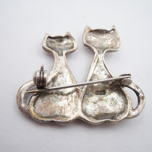 May include: A silver-toned brooch featuring two stylized cat figures side-by-side. The cats have a simple, elegant design with pointed ears and curved tails. The brooch has a pin closure on the back and is marked with "925".
