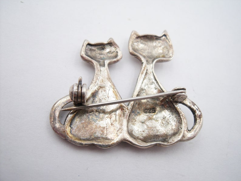 May include: A silver-toned brooch featuring two stylized cat figures side-by-side. The cats have a simple, elegant design with pointed ears and curved tails. The brooch has a pin closure on the back and is marked with "925".