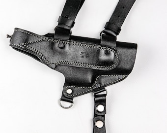 May include: Black leather shoulder holster with adjustable straps and a stitched design. The holster features a molded shape, metal hardware, and a D-ring attachment. The design includes a decorative emblem and is set against a white background.