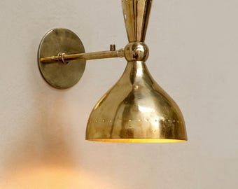 May include: A brass wall sconce with a conical shade and a round backplate. The sconce has a long arm and a decorative top. The shade has small perforations around the bottom. The light is on, emitting a warm glow.