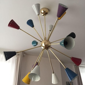 Puede incluir: Una lámpara de araña de estilo retro con una esfera central dorada y múltiples brazos radiantes. Cada brazo sostiene una pantalla cónica de diferentes colores, incluyendo rojo, azul, blanco, amarillo y negro. La lámpara está montada en un techo blanco.