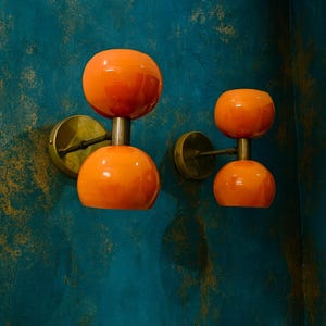 May include: Two orange wall sconces with a double-orb design. Each sconce features a polished brass base and stem, supporting two spherical orange glass shades. The sconces are mounted on a teal wall with gold accents, creating a retro aesthetic.