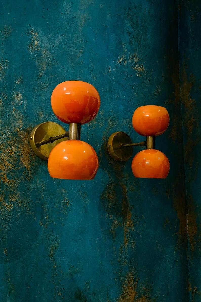 May include: Two orange wall sconces with a double-orb design. Each sconce features a polished brass base and stem, supporting two spherical orange glass shades. The sconces are mounted on a teal wall with gold accents, creating a retro aesthetic.