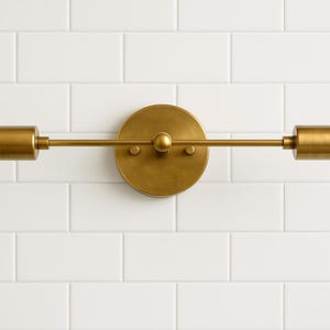 Op de afbeelding: Een messing wandlamp met twee cilindrische lampen die zich uitstrekken vanaf een cirkelvormige basis. De lamp heeft een minimalistisch ontwerp met een warme metalen afwerking en is gemonteerd op een witte betegelde muur.