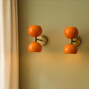 May include: Two orange glass wall sconces with a double-orb design. Each sconce has a brass-colored stem and wall mount. The sconces are mounted on a light green wall, creating a retro, mid-century modern aesthetic.