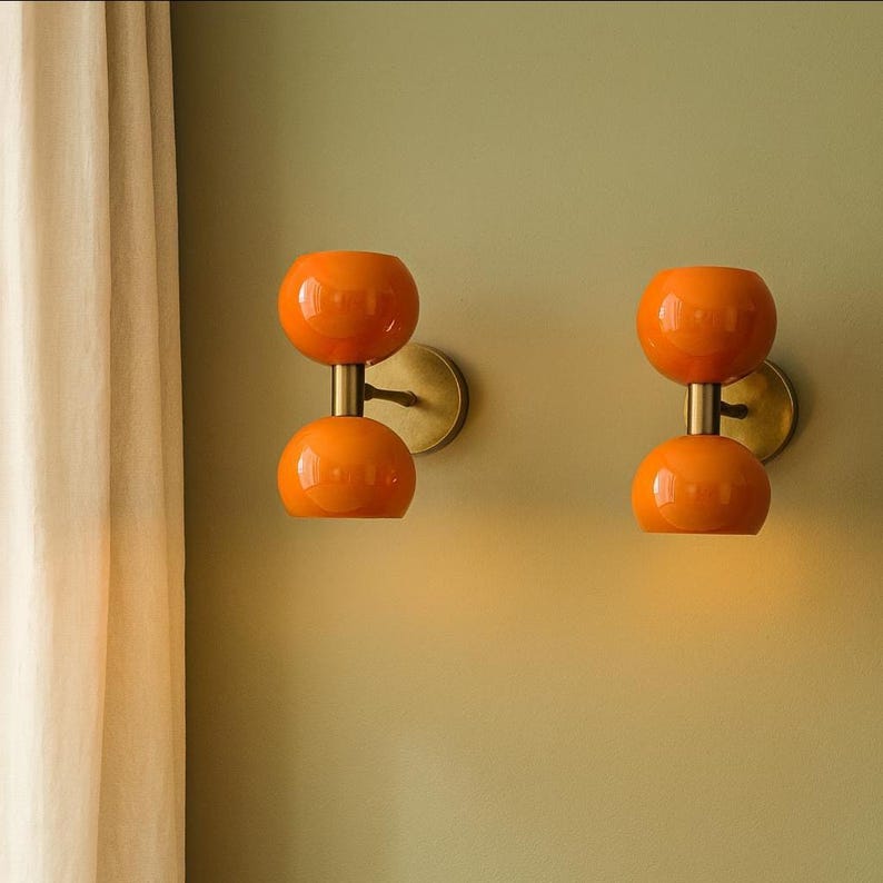 May include: Two orange glass wall sconces with a double-orb design. Each sconce has a brass-colored stem and wall mount. The sconces are mounted on a light green wall, creating a retro, mid-century modern aesthetic.
