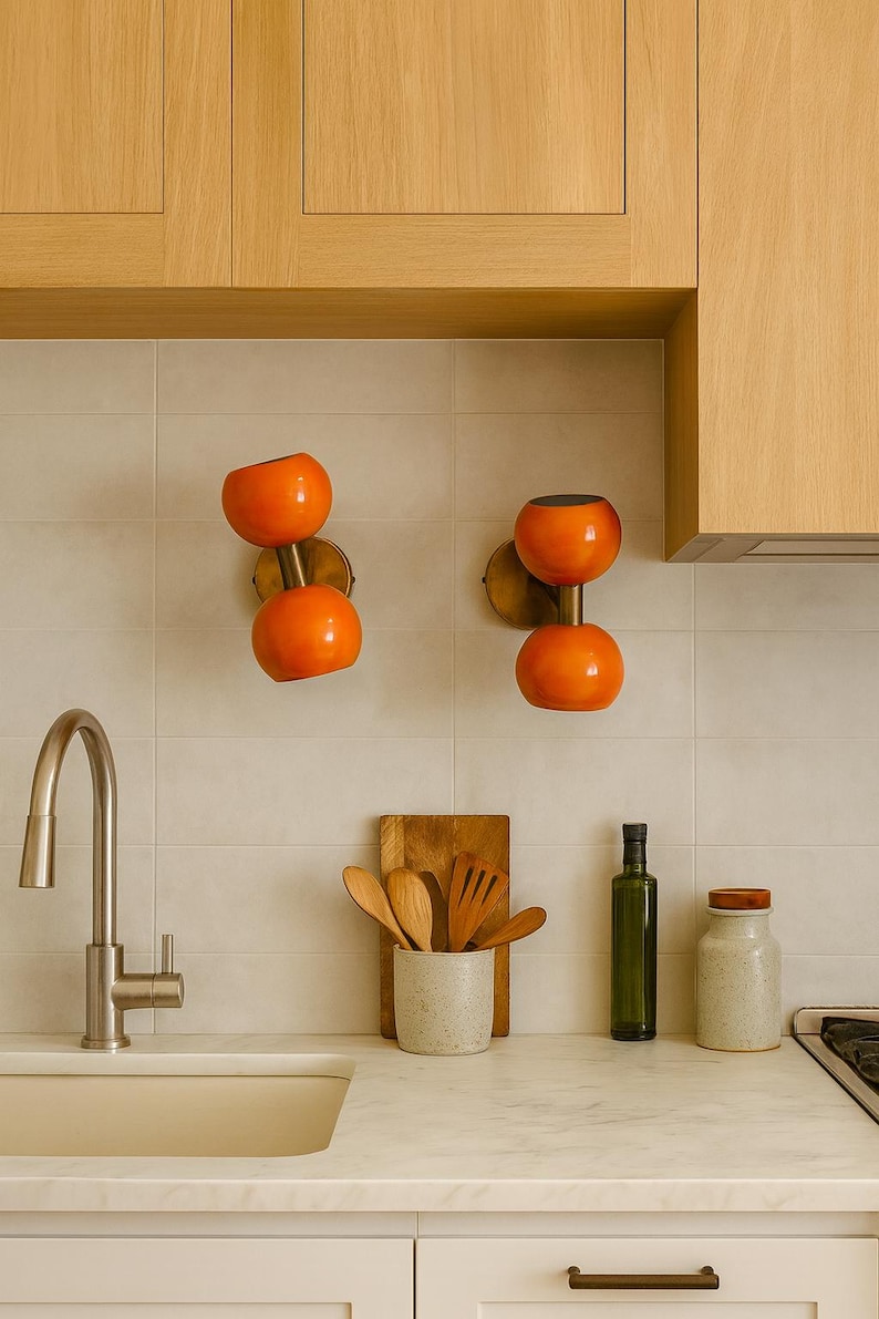 May include: Two orange globe-shaped wall sconces with brass fixtures are mounted on a tiled wall. A kitchen sink, wooden utensils, and a bottle of olive oil are on the countertop. The cabinets are light wood.