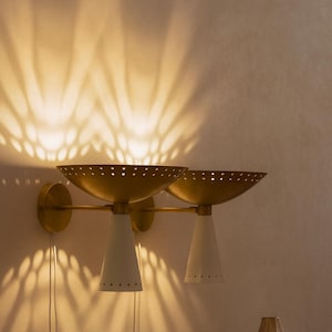 May include: A pair of wall-mounted sconces with a mid-century modern design. The sconces feature gold-colored, bowl-shaped shades with perforations, white cone-shaped bases, and gold-colored mounting hardware. The light casts a patterned shadow on the wall.