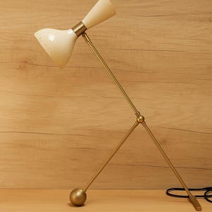 May include: A mid-century modern desk lamp with a cream shade and a brass frame. The lamp features an adjustable design with a cone-shaped shade and a ball-shaped base. The lamp is set on a wooden surface.