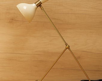 May include: A mid-century modern desk lamp with a cream-colored shade and a brass-colored frame. The lamp has a unique, adjustable design with a cone-shaped shade and a ball-shaped base. The lamp is on a wooden surface.