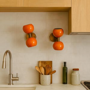 May include: Two orange globe-shaped wall sconces with brass fixtures are mounted on a tiled wall. A kitchen sink, wooden utensils, and a countertop with a marble effect are also visible. The kitchen has light wood cabinets.