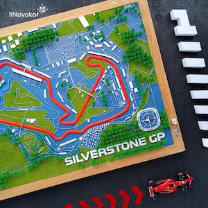 Könnte beinhalten: Ein gerahmter 3D-Plan der Silverstone GP-Rennstrecke mit roter Streckenführung auf einer blau-grünen Landschaft. Die Worte "SILVERSTONE GP" sind zu sehen. Ein kleines rotes Modellrennauto befindet sich darunter.