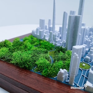 This image showcases a detailed 3D printed miniature map of Manhattan's Central Park, featuring lush green trees and pathways, set against a white cityscape of tall buildings. The model is mounted on a sleek wooden base, highlighting the contrast between the natural green spaces and urban architecture. The design captures the essence of Central Park within the city skyline, making it a modern wall art piece and a perfect Manhattan-themed gift, emphasizing craftsmanship and city pride.