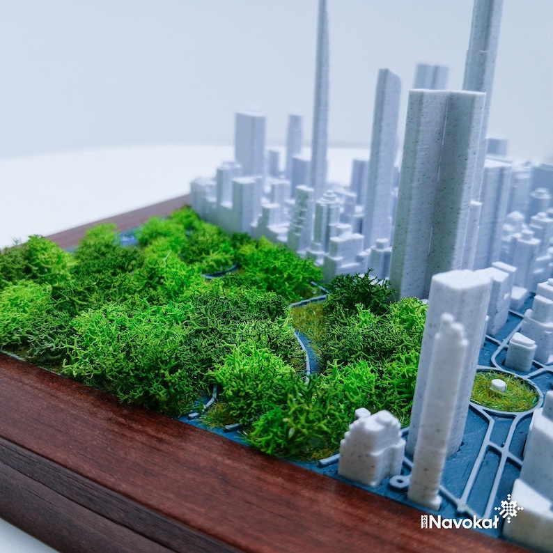 This image showcases a detailed 3D printed miniature map of Manhattan's Central Park, featuring lush green trees and pathways, set against a white cityscape of tall buildings. The model is mounted on a sleek wooden base, highlighting the contrast between the natural green spaces and urban architecture. The design captures the essence of Central Park within the city skyline, making it a modern wall art piece and a perfect Manhattan-themed gift, emphasizing craftsmanship and city pride.