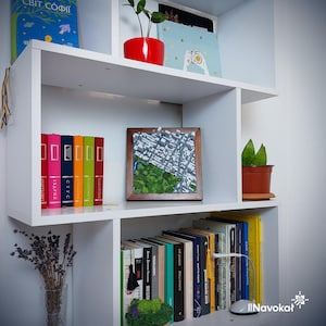 This photo displays a modern white bookshelf with a framed 3D map of Central Park in Manhattan, New York City. The map features detailed white buildings, green trees, and pathways, showcasing a miniature cityscape. The shelf is decorated with colorful books, a potted plant, and artwork, creating an appealing background that highlights the stylish, wooden-framed wall art. The setting emphasizes the artwork's contemporary design and urban theme as a unique gift or decor piece.