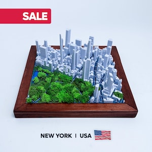 This photo features a detailed 3D miniature map of Manhattan's Central Park, showcasing lush green trees and pathways surrounded by a white cityscape of tall buildings, mounted on a sleek wooden base. The map highlights New York City's iconic urban and green spaces, making it a stylish modern wall art piece. The wooden frame adds a touch of elegance, perfect as a Manhattan-themed gift or unique home decor for city lovers.