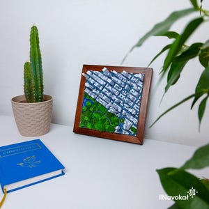 This image features a framed 3D miniature map of Central Park in Manhattan, New York City, displayed on a white surface. The map showcases detailed white buildings and lush green trees representing the park, with a circular pathway and water feature. The wooden frame adds a modern touch to the artwork. Surrounding the frame are a potted cactus, a green plant branch, and a blue book, creating an elegant and stylish display of this Manhattan-themed wall art and gift.