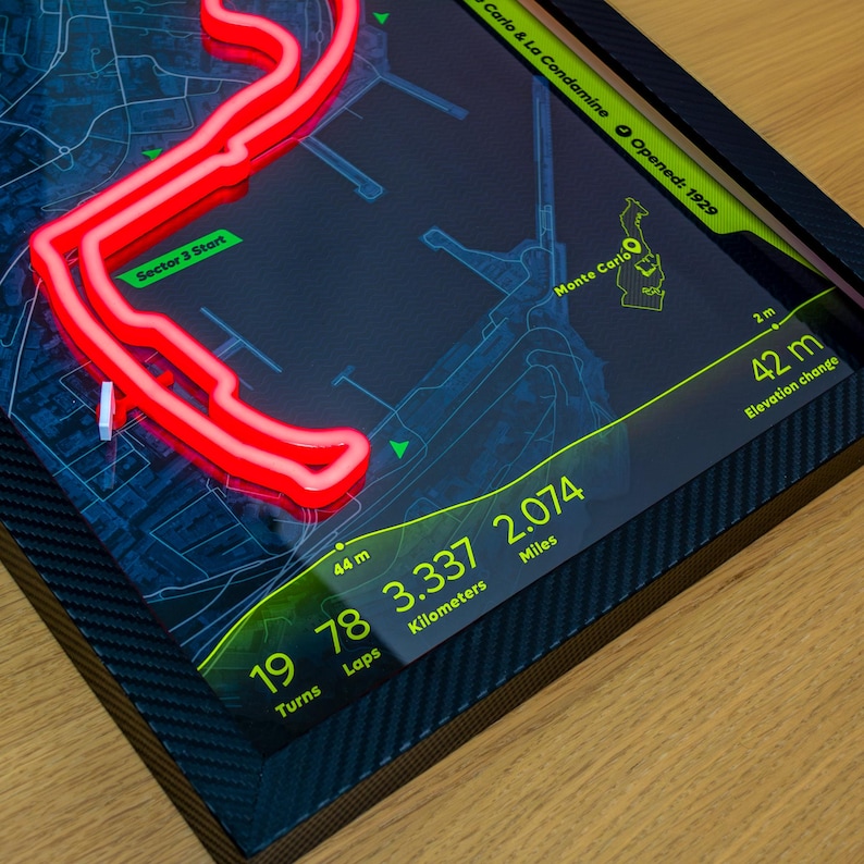 May include: A framed wall art piece featuring a glowing red neon outline of the Monte Carlo race track on a dark blue map background. The display includes text details such as 19 turns, 78 laps, 3.337 kilometers, 2.074 miles, and 42 m elevation change.