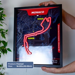 Framed 16×20-inch Monaco LED neon racetrack map wall art inspired by Formula 1, featuring the iconic Monaco Grand Prix circuit glowing in bright red over a dark map background. The black carbon fiber-style frame is held against a white wall, with circuit details, sector markers, and Monte Carlo map graphics visible. A unique motorsport gift and stylish racing-themed home decor piece.