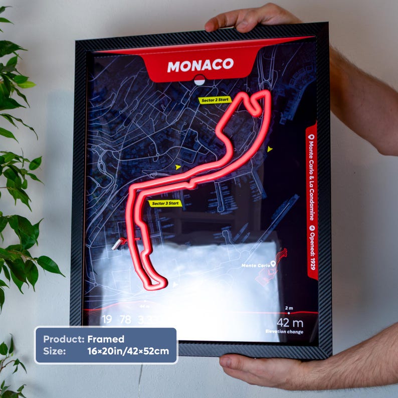 Framed 16×20-inch Monaco LED neon racetrack map wall art inspired by Formula 1, featuring the iconic Monaco Grand Prix circuit glowing in bright red over a dark map background. The black carbon fiber-style frame is held against a white wall, with circuit details, sector markers, and Monte Carlo map graphics visible. A unique motorsport gift and stylish racing-themed home decor piece.