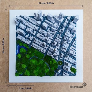 This image shows a 3D printed miniature map of Central Park in Manhattan, New York City, framed with a modern wood border. The map features detailed white buildings, green trees, and pathways, highlighting the iconic park amid the cityscape. The artwork is placed on a wooden surface with decorative items like books and plants around it, emphasizing its stylish design and urban theme, making it a perfect modern wall art or gift representing Manhattan.