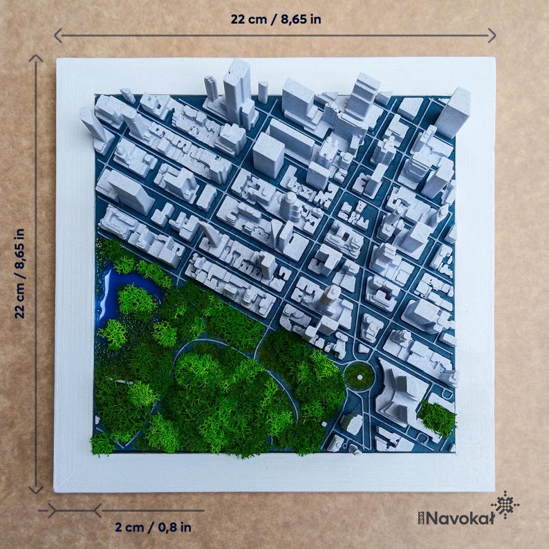 This image shows a 3D printed miniature map of Central Park in Manhattan, New York City, framed with a modern wood border. The map features detailed white buildings, green trees, and pathways, highlighting the iconic park amid the cityscape. The artwork is placed on a wooden surface with decorative items like books and plants around it, emphasizing its stylish design and urban theme, making it a perfect modern wall art or gift representing Manhattan.