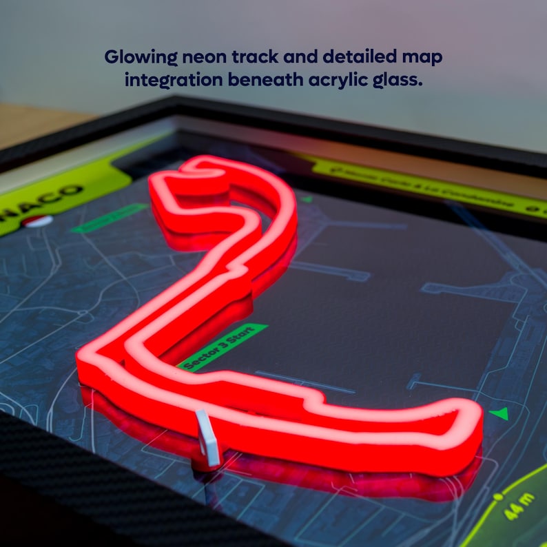 May include: A framed wall art piece featuring a glowing red neon race track outline of Monaco set against a detailed dark map background under acrylic glass. The text "Glowing neon track and detailed map integration beneath acrylic glass" is visible.