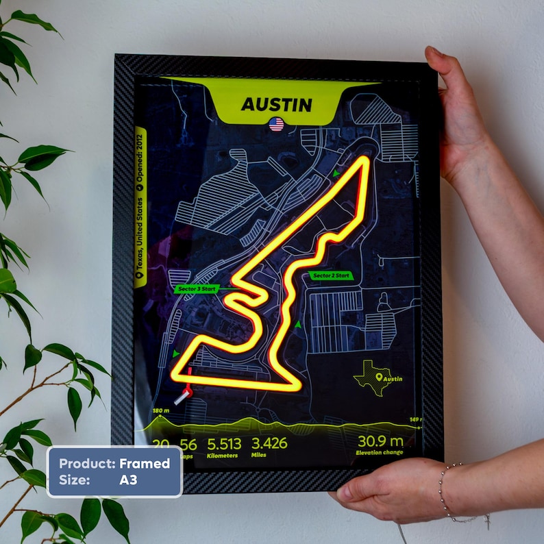 Framed A3 Austin LED neon racetrack map wall art inspired by Formula 1, featuring the Circuit of the Americas track glowing in orange and yellow over a dark map background. The black carbon fiber-style frame is held by hands against a white wall, with circuit statistics, sector markers, and a Texas outline visible, making a unique motorsport gift and home decor piece.