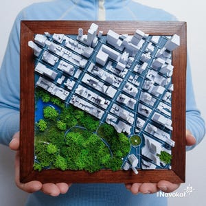 This photo shows a person holding a framed 3D miniature map of Central Park in Manhattan, New York City. The map features white 3D printed buildings representing the city skyline, with a detailed green landscape of trees and pathways in the park area. The wooden frame enhances the modern aesthetic, making it a stylish wall art piece. The intricate design highlights the contrast between urban structures and natural green space, perfect as a Manhattan-themed gift or decor.