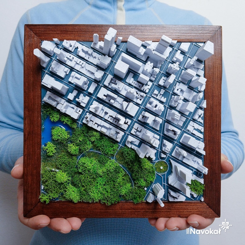 This photo shows a person holding a framed 3D miniature map of Central Park in Manhattan, New York City. The map features white 3D printed buildings representing the city skyline, with a detailed green landscape of trees and pathways in the park area. The wooden frame enhances the modern aesthetic, making it a stylish wall art piece. The intricate design highlights the contrast between urban structures and natural green space, perfect as a Manhattan-themed gift or decor.