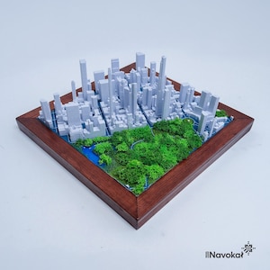 This photo features a detailed 3D miniature map of Manhattan's Central Park, showcasing lush green trees and pathways surrounded by a white cityscape of tall buildings, mounted on a sleek wooden base. The map highlights New York City's iconic urban and green spaces, making it a stylish modern wall art piece. The wooden frame adds a touch of elegance, perfect as a Manhattan-themed gift or unique home decor for city lovers.
