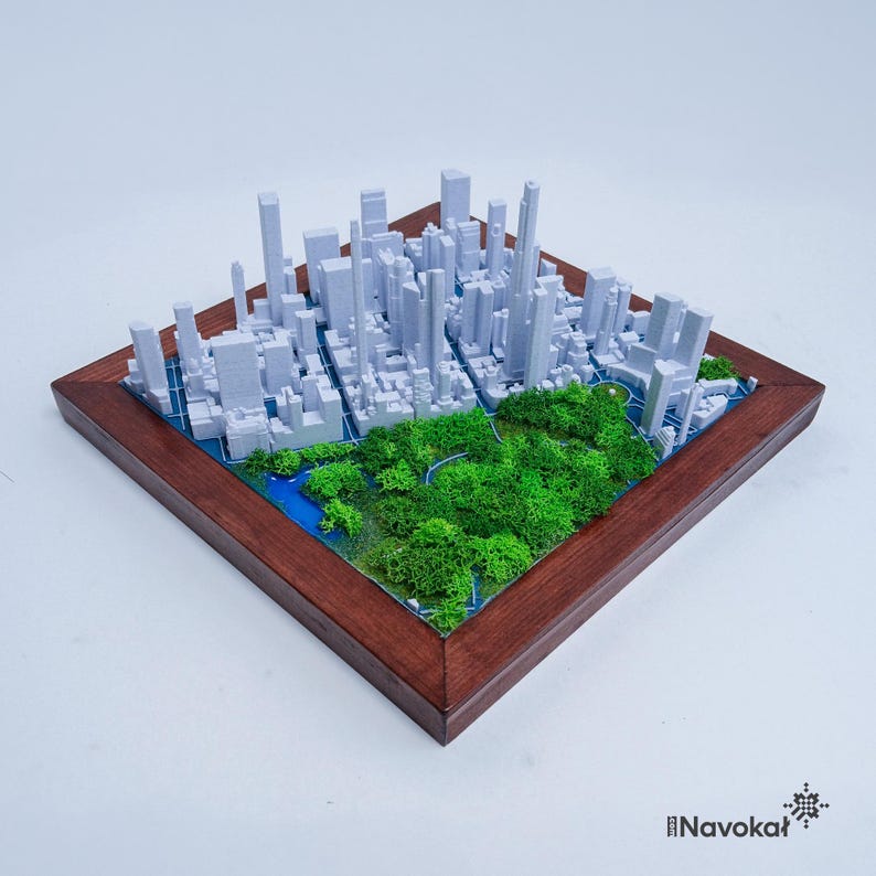 This photo features a detailed 3D miniature map of Manhattan's Central Park, showcasing lush green trees and pathways surrounded by a white cityscape of tall buildings, mounted on a sleek wooden base. The map highlights New York City's iconic urban and green spaces, making it a stylish modern wall art piece. The wooden frame adds a touch of elegance, perfect as a Manhattan-themed gift or unique home decor for city lovers.