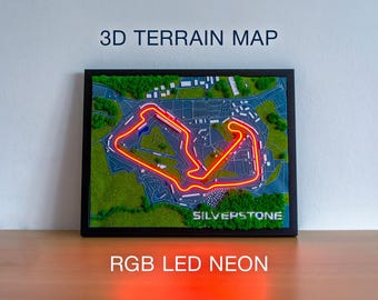 Könnte beinhalten: Eine gerahmte 3D-Geländekarte der Rennstrecke Silverstone mit einer neonbeleuchteten Streckenkontur. Die Karte zeigt grüne, blaue und weiße Elemente mit dem Text "3D TERRAIN MAP" und "SILVERSTONE". Der Text unten lautet "RGB LED NEON".