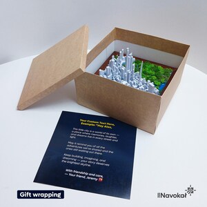This image displays the inside of a brown cardboard gift box containing a 3D printed miniature map of Central Park in Manhattan, New York City, with detailed white buildings and lush green trees. The box is open, revealing the artwork, and a personalized message card with customizable text is placed beneath it. The card includes an example message and a note about the gift’s sentimental value. The setup highlights the modern, stylish presentation perfect for gifting or decorating a Manhattan.