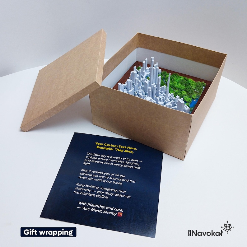 This image displays the inside of a brown cardboard gift box containing a 3D printed miniature map of Central Park in Manhattan, New York City, with detailed white buildings and lush green trees. The box is open, revealing the artwork, and a personalized message card with customizable text is placed beneath it. The card includes an example message and a note about the gift’s sentimental value. The setup highlights the modern, stylish presentation perfect for gifting or decorating a Manhattan.