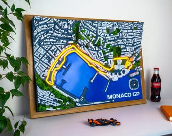 Könnte beinhalten: Ein 3D-Wandkunstwerk, das den Monaco GP-Kurs mit einer leuchtend gelben Strecke, einem blauen Hafen und weißen Stadtgebäuden in einem Holzrahmen darstellt. Der Text MONACO GP ist unten rechts auf dem blauen Hafenbereich in Weiß aufgedruckt.