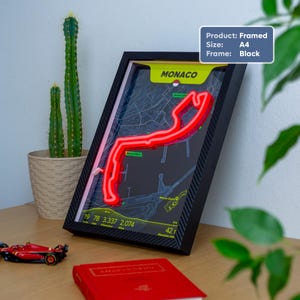 May include: A black A4 framed wall art piece featuring a neon red Monaco Grand Prix circuit map on a dark background. The design includes the text MONACO, race statistics, and sector markers. The frame has a carbon fiber texture finish.