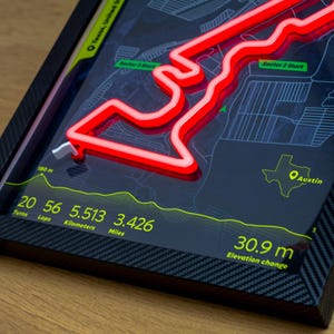 Close-up of an Austin Formula 1 Circuit of the Americas LED wall art poster, showing a glowing red neon track layout mounted on a dark map background. Fine details include sector start markers, an elevation profile, and track stats (20 turns, 5.513 km / 3.426 miles, 30.9 m elevation change), with a carbon-fiber-style frame, celebrating the United States Grand Prix and Charles Leclerc’s fastest lap.