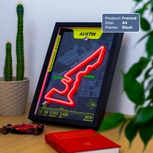 Framed Austin Neon F1 Track Poster featuring the United States Grand Prix circuit in Austin, Texas. The A4 wall art is displayed in a black frame with a bright red LED-style track outline representing Charles Leclerc’s Ferrari fastest lap. The design includes a dark map background, circuit sector markers, track statistics, and elevation data. Shown standing on a wooden desk beside a model Formula 1 car, a red book, and potted plants in a modern home setting.